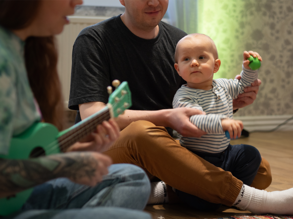 Poika soittaa kahta rytmimunaa isän avustuksella. Etualalla muskariopettaja Simonette Ohra-aho soittaa ukulelea ja laulaa.